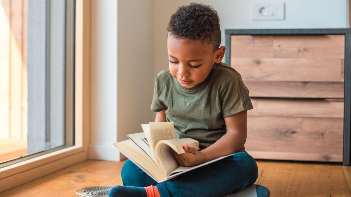 Young child flicking through an adult book in his home. He is wearing a khaki green T-shirt and jeans and has short black hair.