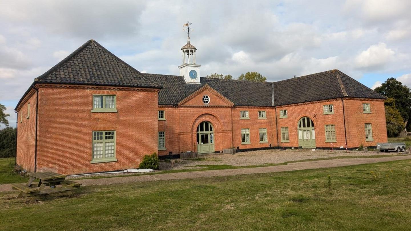 A large redbrick building in the shape of a horse shoe with with sage green window frames and a clock tower on top