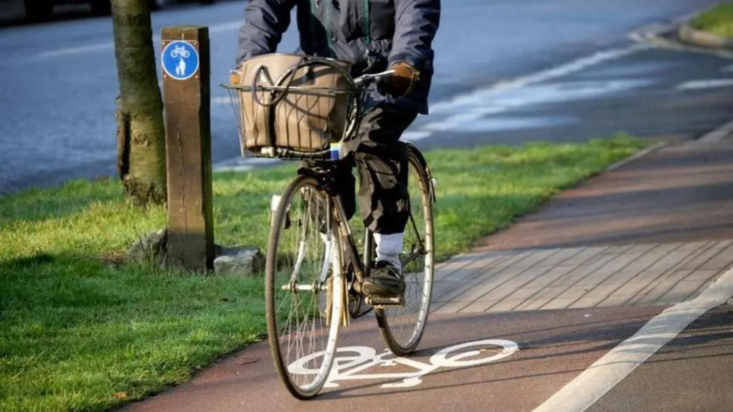 A person on a bicycle, riding along a cycle path. There is a grass verge to their right, which runs along the main road.