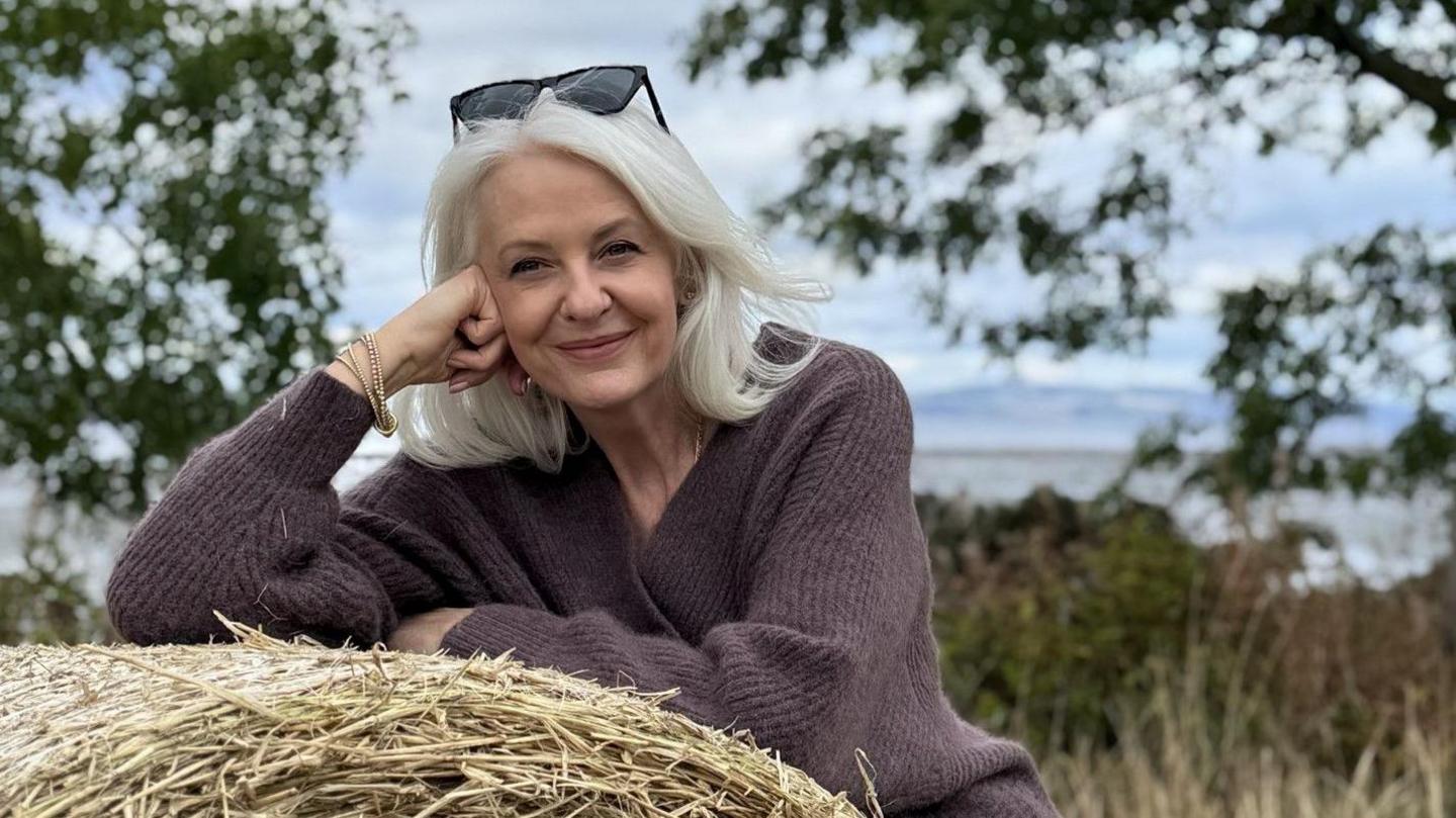 Valerie is outside, in a field, leaning on a bale of hay. She's wearing a purple/brown knit jumper and has black sunglasses perched on top of her head. She's smiling.
