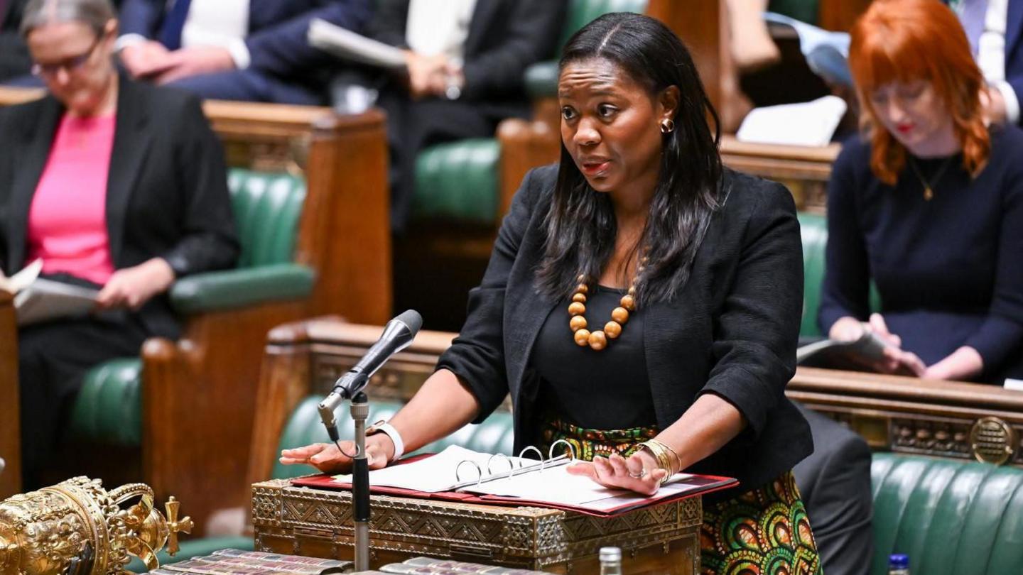 A woman in a black jacket and top with a colourful necklace and skirt stands at the despatch box in the House of Commons. Some female MPs are visible on the benches behind her.