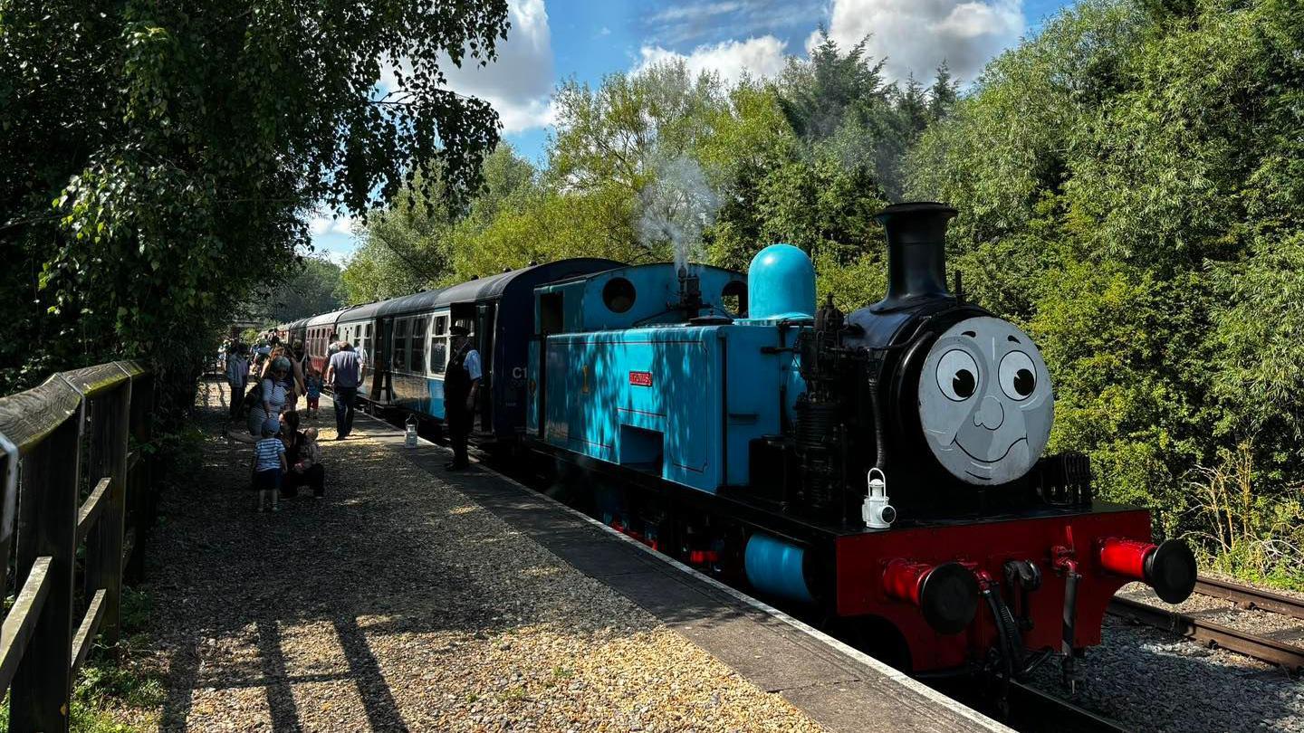 Thomas the tank engine- blue steam train with a smiley face in front- at the platform with members of the public queuing up to board it.