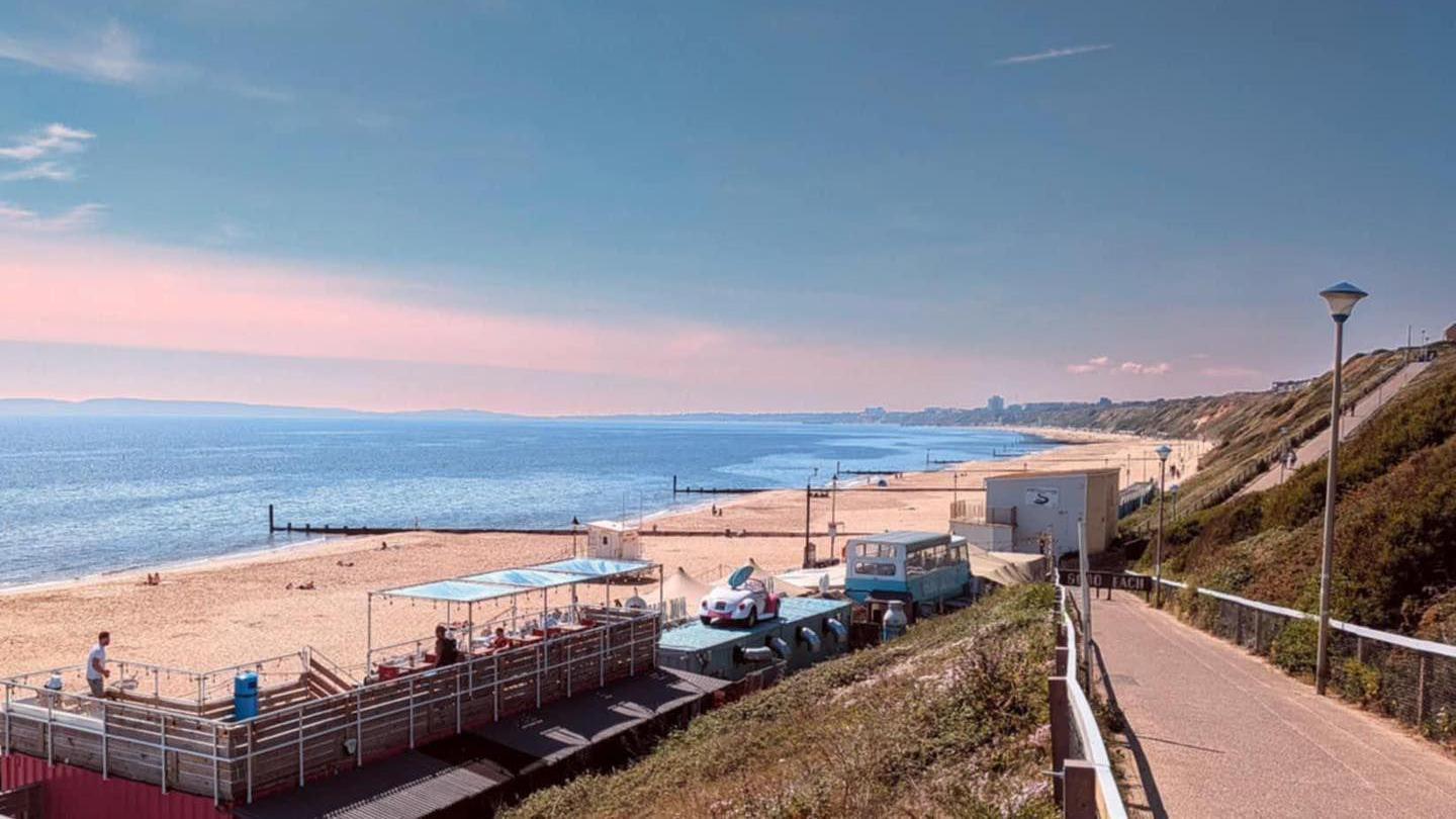 View from the zig-zag footpath path from the cliff top, looking down towards Sobo Beach bar and the long sandy beach of Southbourne and Bournemouth. The beach bar is made up of a converted double decker bus, a smaller building with air vents, and a large open decking area.