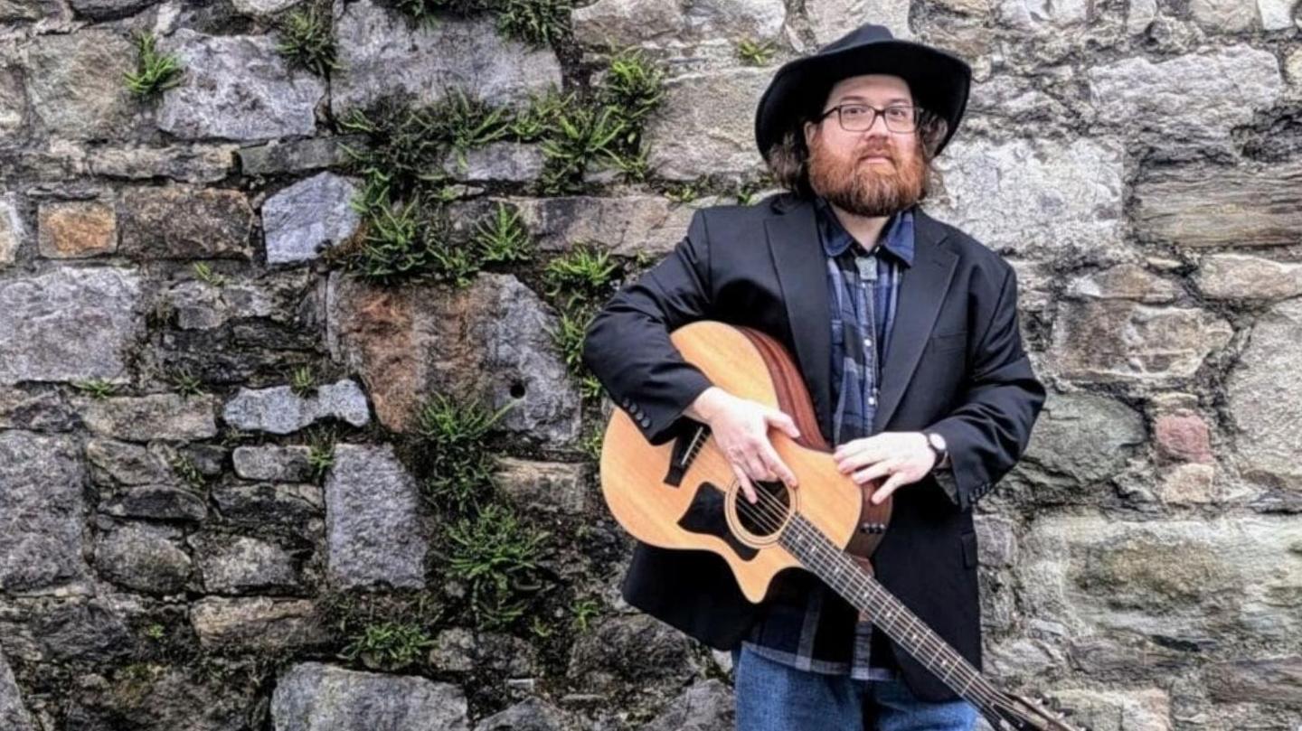 Llun o Joshua Gardner yn sefyll o flaen wal gerrig. Mae'n gwisgo het cowboi ac yn dal gitar.