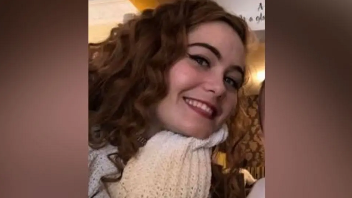 A smiling Courtney Mitchell looks directly at the camera with her head tilted to her left. She is a young woman with long, slightly curly dark brown/ginger hair and is wearing a white scarf.
