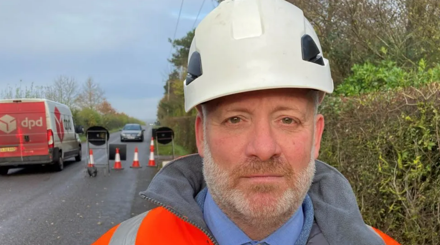 Darren Clark wearing a white hard hat with a road with cones behind him and a DPD van passing