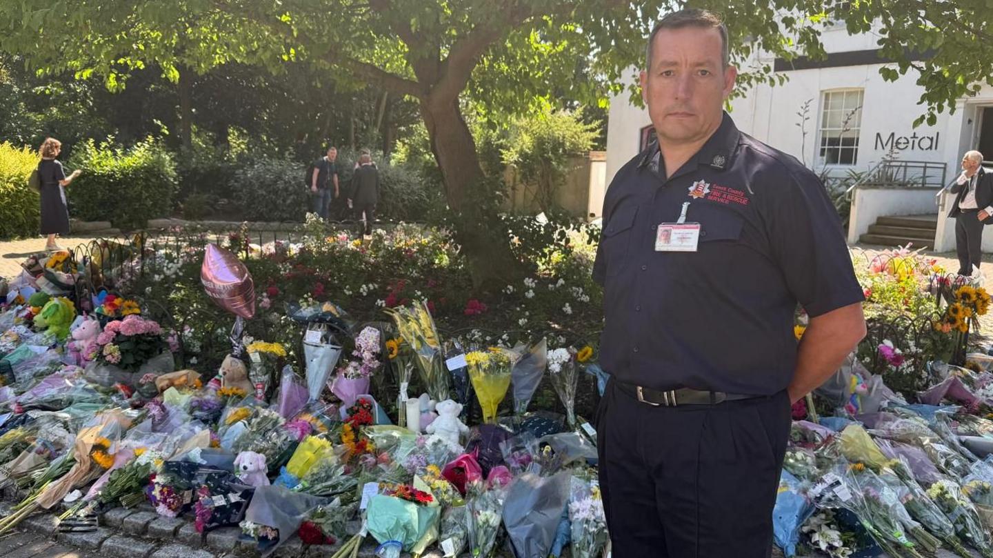 A man standing in his uniform with a background of flowers spread across a tree. Teddy bears are also being left. The man has a sombre expression on his face and he has his hands behind his back.
