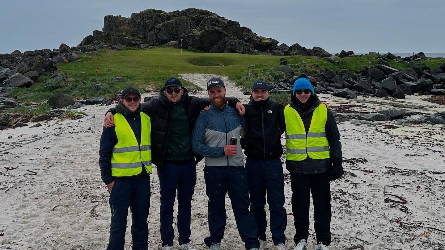 A group of four men are stood on shallow snow with a green golf course behind them. In the middle is Isaac, who is smiling. 
