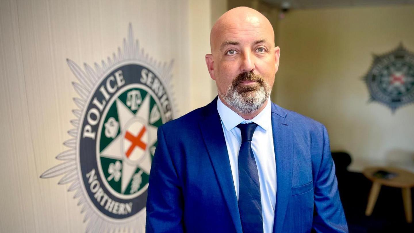 Man with bald head and grey beard. He is wearing a blue suit and tie with white shirt, standing in front of a PSNI badge on a wall