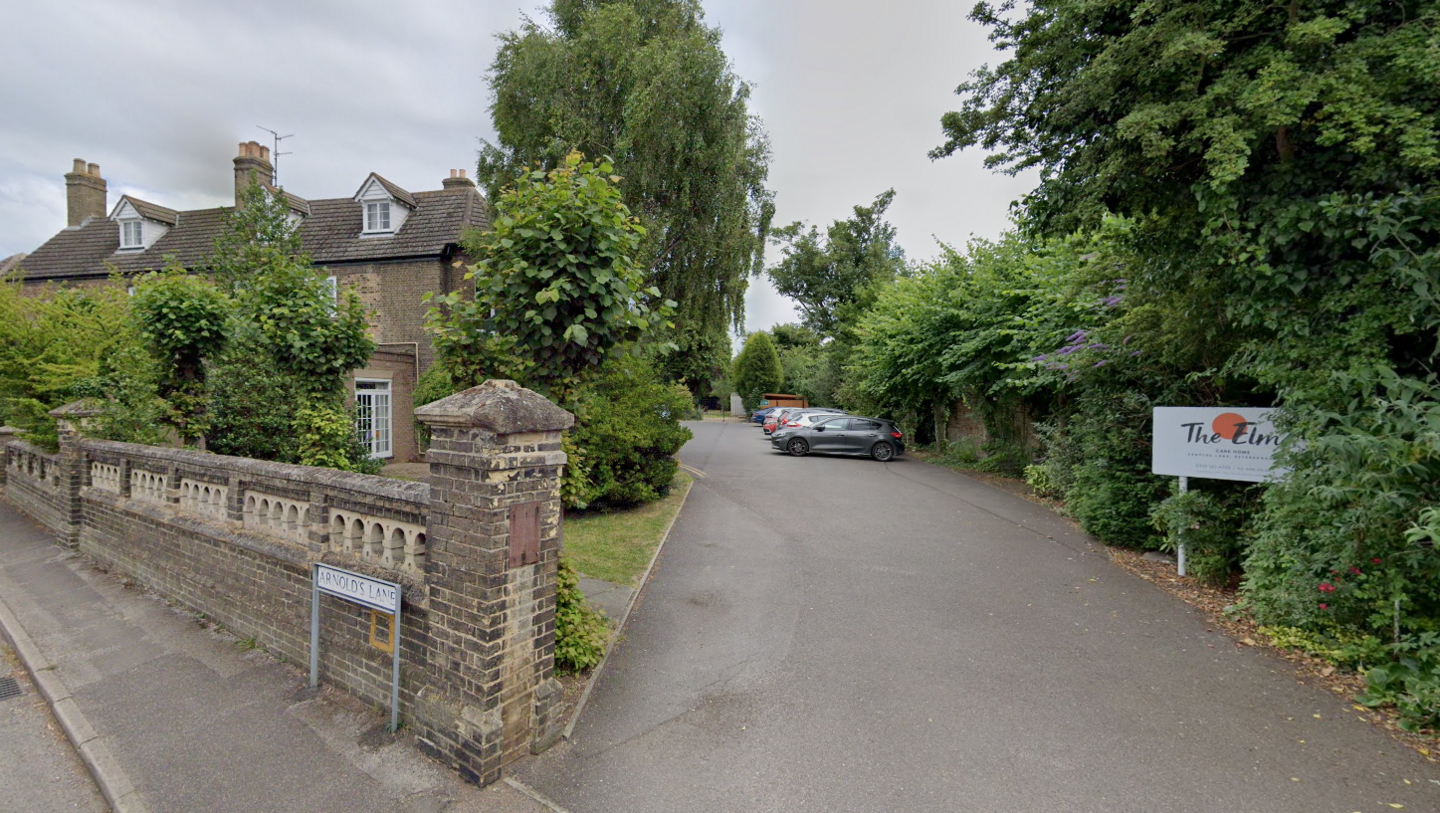 Street view of the former care home, pictured in 2022. It is surrounded by a wall with large trees and greenery around its edges. There is a long driveway to the front of the property and a white sign to the left which reads 'The Elms'.