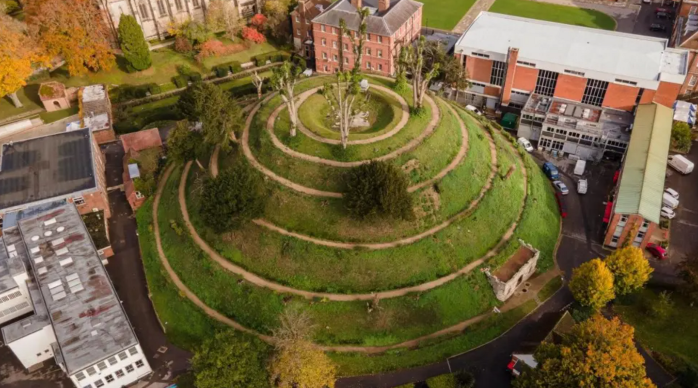 A drone shot of mound, showing the spiral pathway all the way round to the top. It is mostly covered in grass, but there is the odd tree and a small grotto area at the side. It is surrounded by school buildings, mostly red brick and several storeys high.