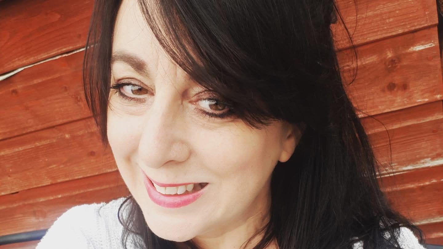 A close up image of an older women with pale skin. She has black hair with a side fringe swooping over her forehead. She is wearing a copper eyeshadow with black eyeliner and a light pink lipstick.