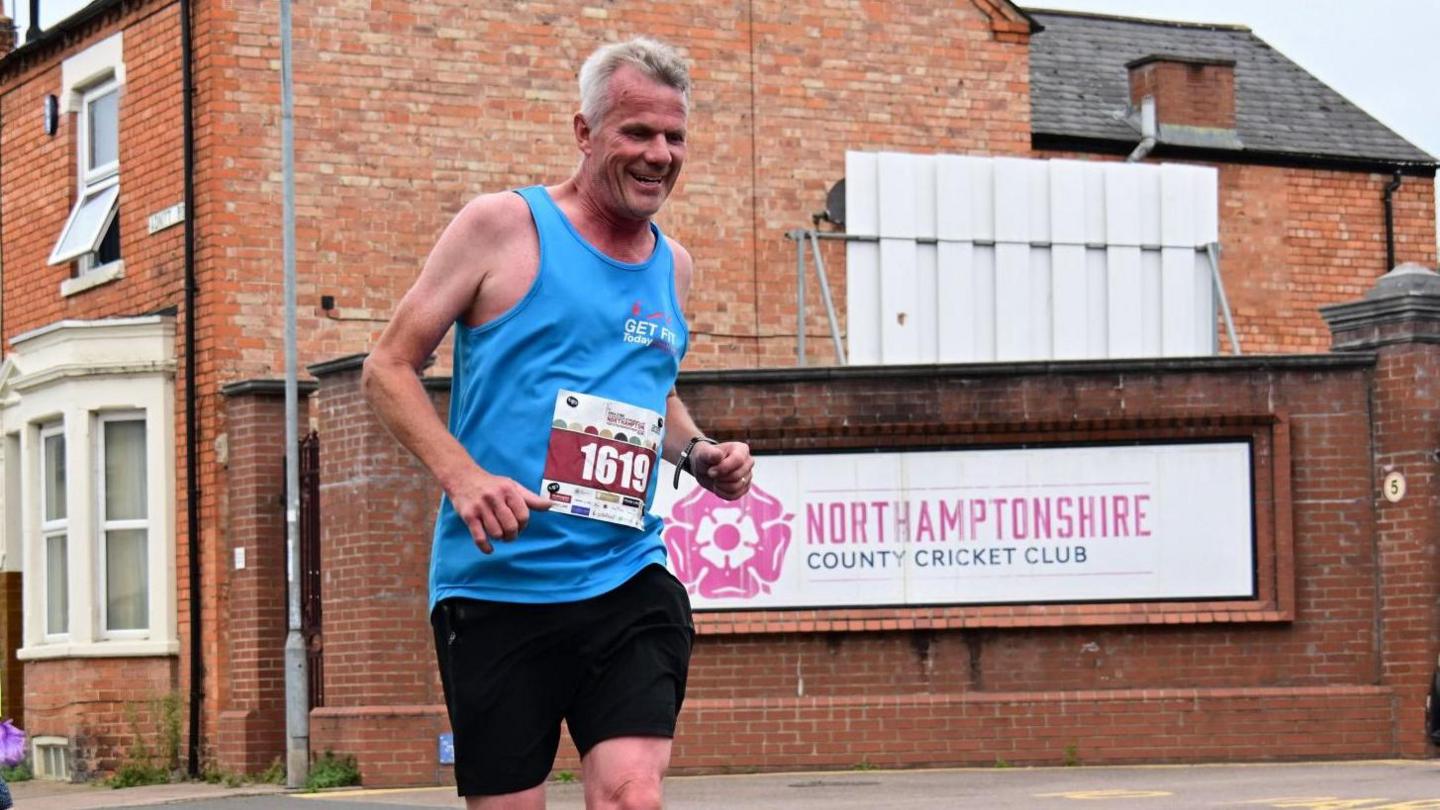 A male runner in a blue vest runs past a sign for Northamptonshire County Cricket Club's ground.