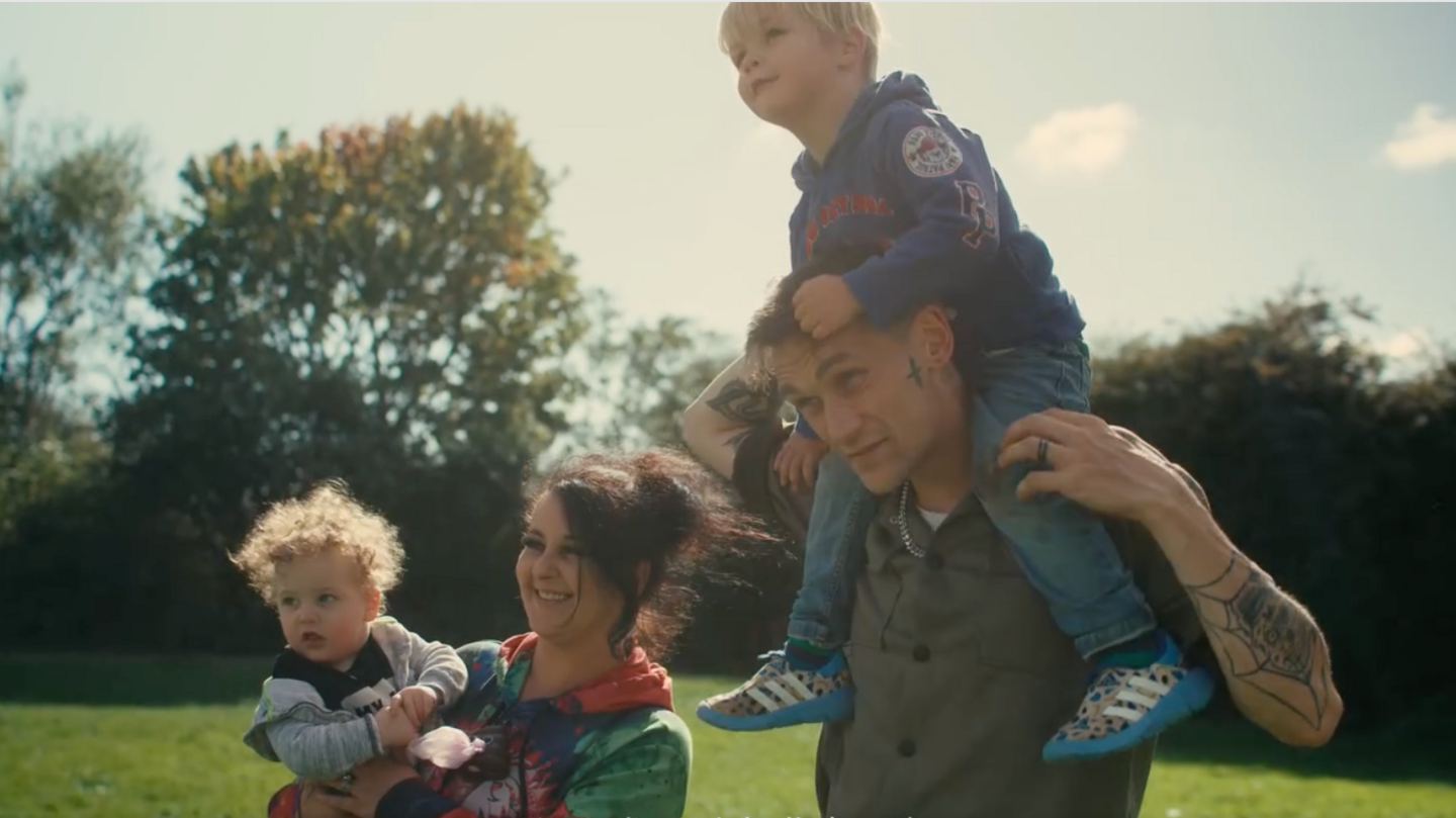 Rob with his family. He is wearing a green shirt and has a child sat on his shoulders. His partner is stood next to him holding their other child in her arms.