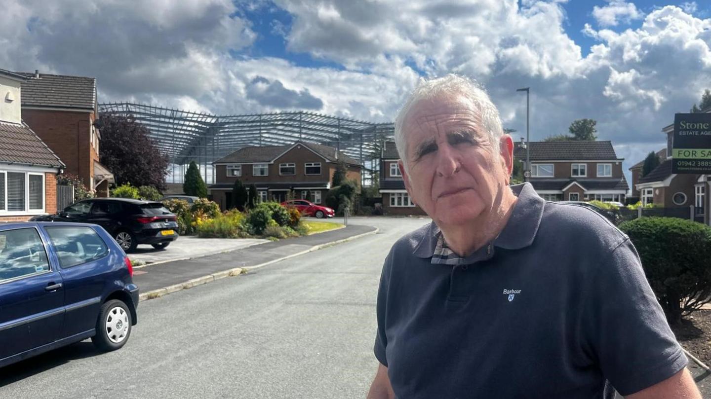 A man in a blue polo shirt stands in a residential street. The metal structure of a large warehouse is behind the houses.