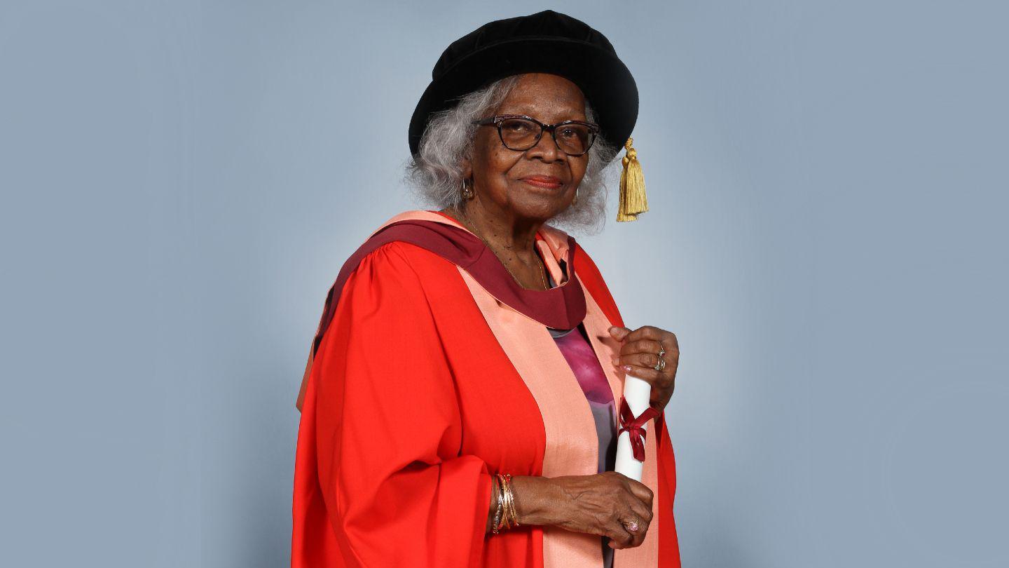 Barbara Dettering wearing her red doctorate robes and holding her honorary graduation scroll, which is white with a red ribbon tied in a bow in the middle. She is wearing a black graduation cap with a gold tassel, and standing against a blue-grey background