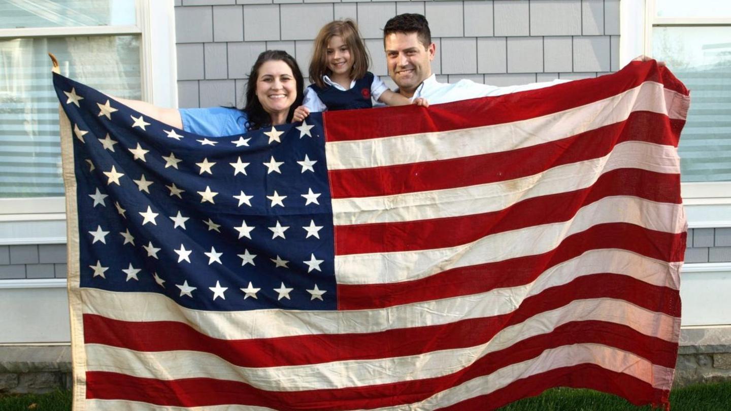 A family of three hold up a stars and stripes flag while smiling outside their home.