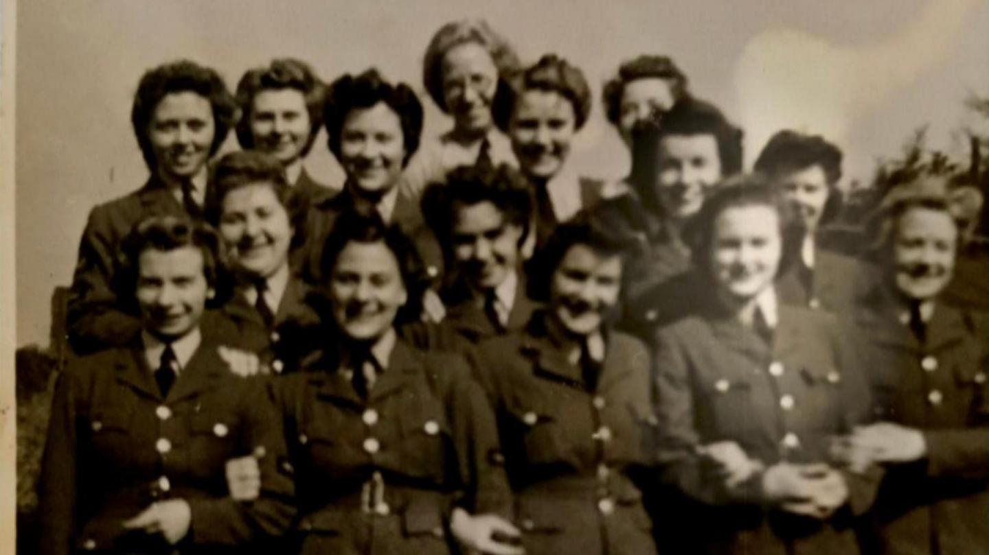 Cynthia Evans is in a black and white photograph with 14 of her female RAF colleagues. They are all wearing their RAF uniforms and are smiling at the camera.
