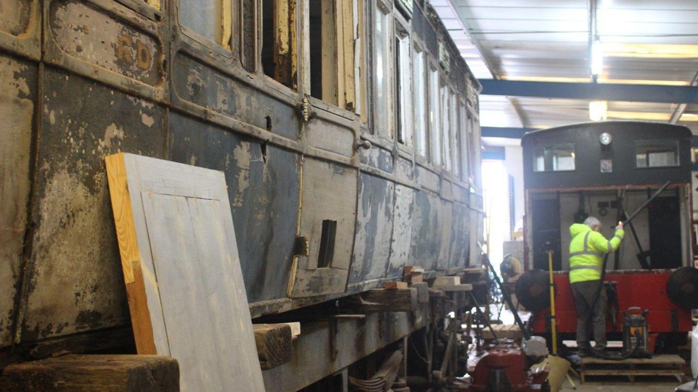 A warehouse containing a dilapidated railway carriage with Third written on the doors. A man in a yellow high-vis coat can be seen working on a brake van in the background.