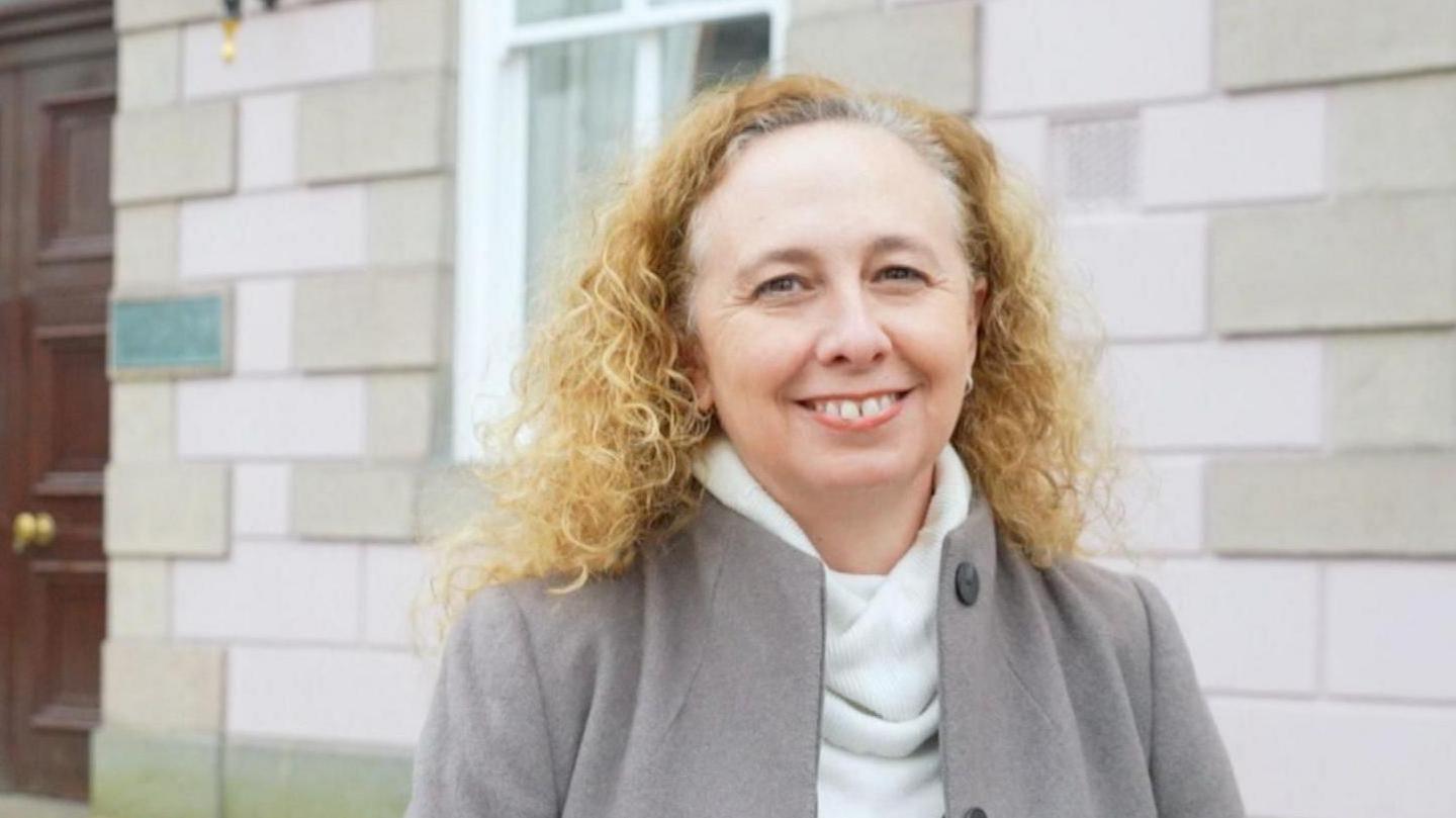 Jersey deputy Inna Gardiner has a broad smile while standing outside a government building. She is wearing a grey goat, white turtle neck jumper and has long curly blonde hair.