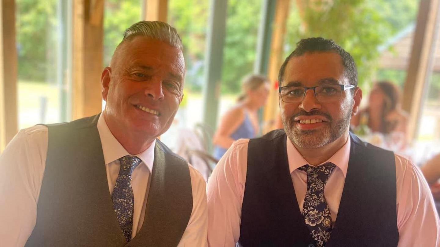 Dayon Kingsland, a mixed-race man wearing a dark-coloured waistcoat and blue floral tie sitting next to Kirk Godber, a white man wearing a grey waistcoat and blue floral tie with a white shirt. Both men are smiling. 