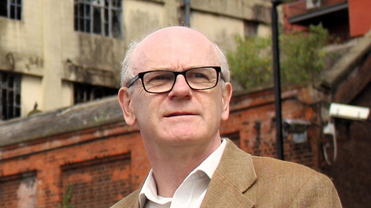 Neil MacDonald looks away from the camera. He is largely bald and is wearing a brown jacket with a white shirt underneath.