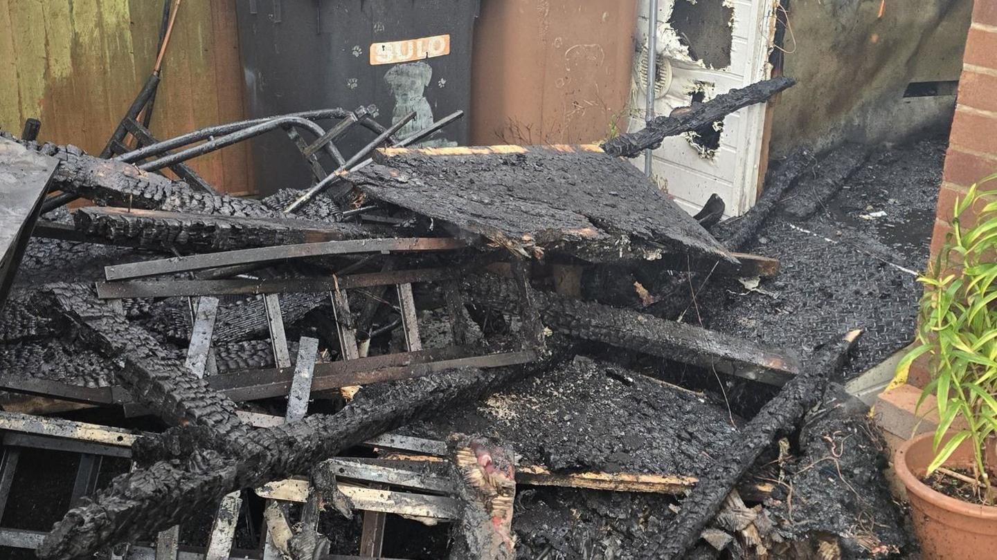 A burnt-down shed reduced to a pile of charred wooden planks on the ground. Next to it is a wooden fence and a redbrick house. Within the heap of remains there are some metal structures - a ladder and table and chair legs can be seen poking out. These have also been blackened by the fire.
