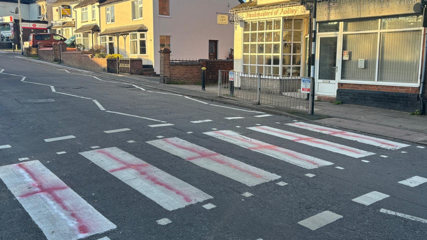 Six white stripes on the crossing that are visible in the photo have been painted with a red cross to look like the England flag. Shops and houses can be seen on the right hand-side of the photo.