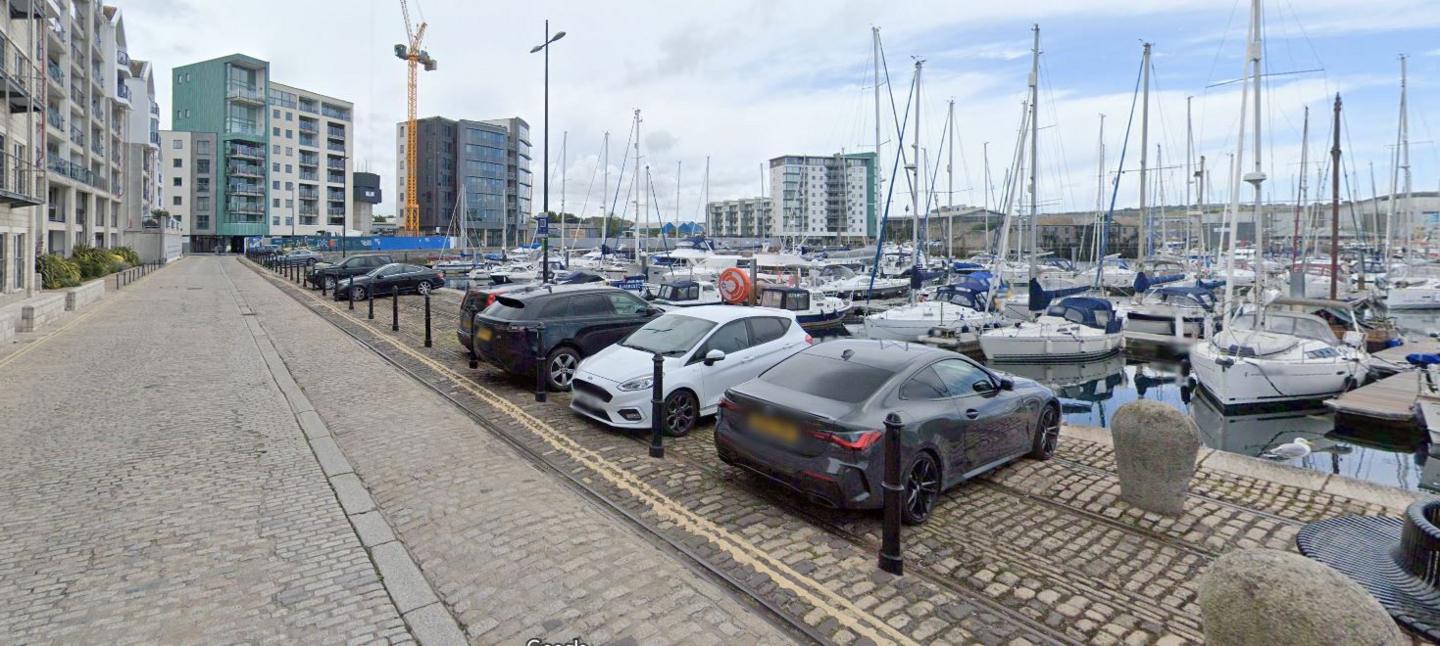 North Quay in Plymouth, with a road to the left, cars parked in the middle and the quay seen to the right, with boats moored in the harbour. 