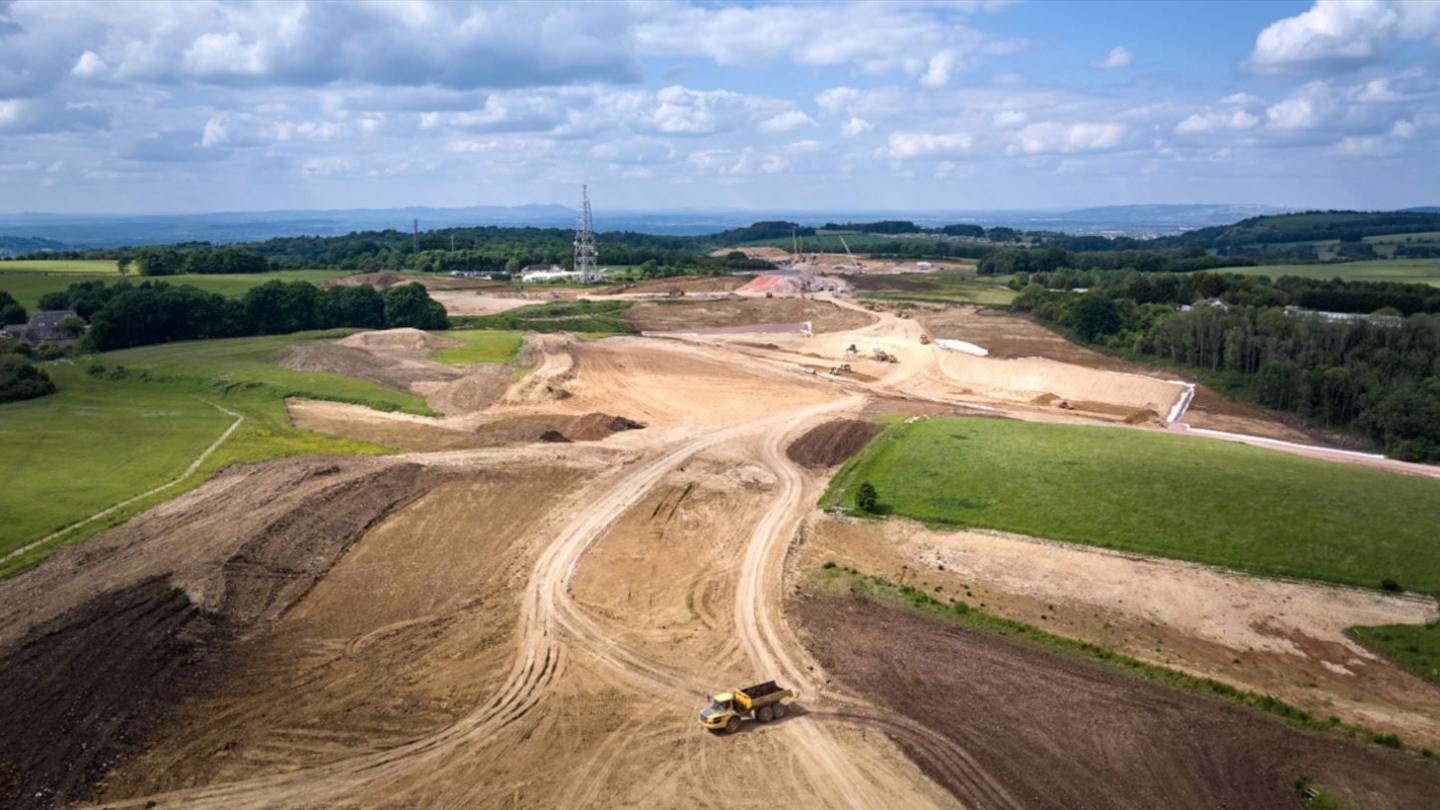 An aerial image showing the progress on the A417 Missing Link scheme. It is a large sprawling patch of land that has been levelled out, surrounded by fields and trees. In the foreground there is a digger truck with a bed full of dirt.