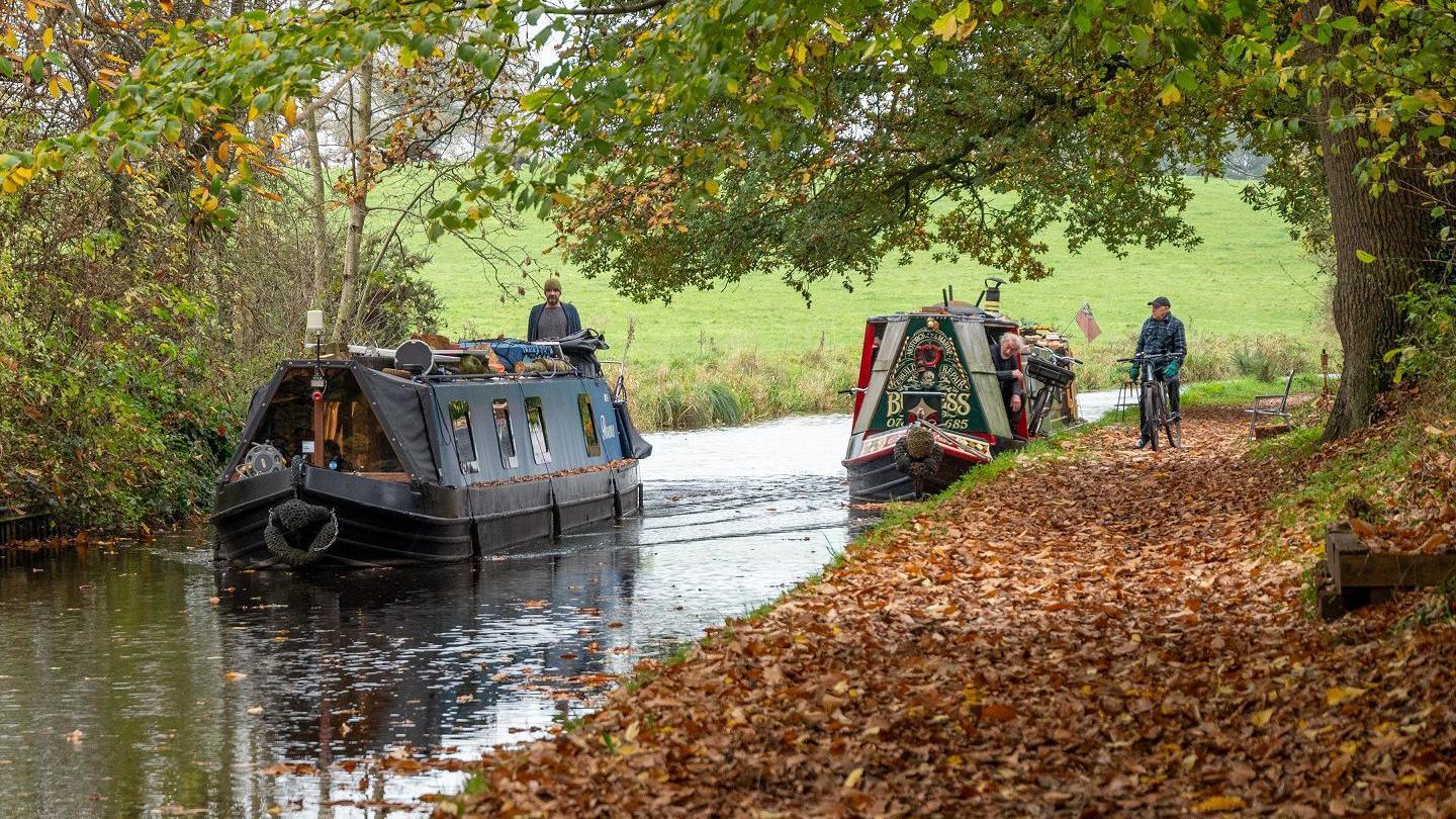 Two narrowboats come towards the camera on a waterway. Alongside the waterway is a path covered in brown leaves with a cyclist visible in a dark outfit. The cyclist stands astride his bike as he looks at the boats going past. The closer of the two boats is blue and black with a man in a woollen green hat at the tiller while the other, further away, has red, green and pale yellow markings. Behind them is a field in the distance with green grass.