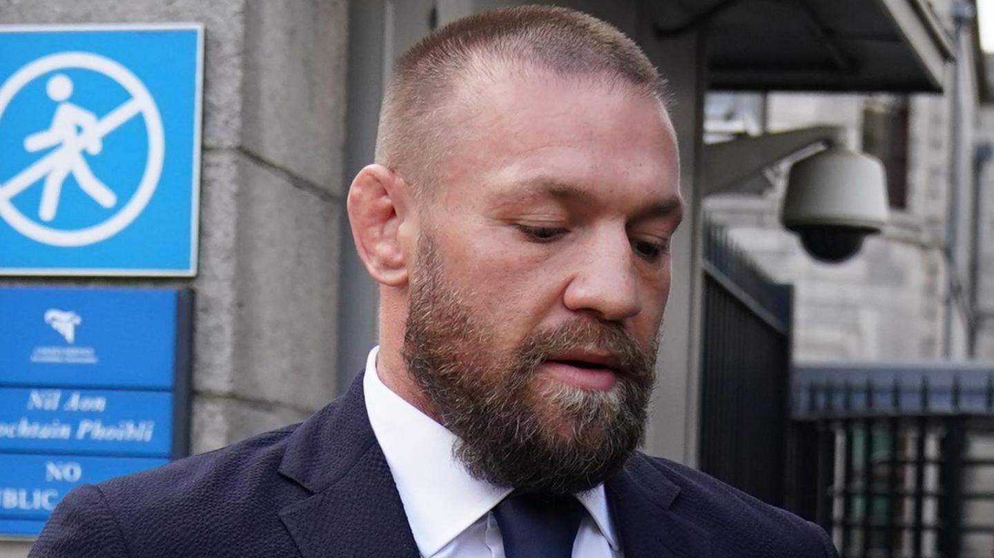 Conor McGregor outside the High Court in Dublin.  He has a beard, shaved hair and is wearing a navy suit and a white shirt.