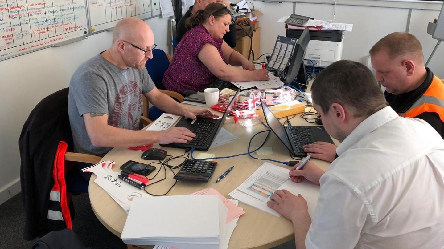 Staff at Norsk Hydro crowd around a small desk. Two are working with pens and paper, another is on a laptop.