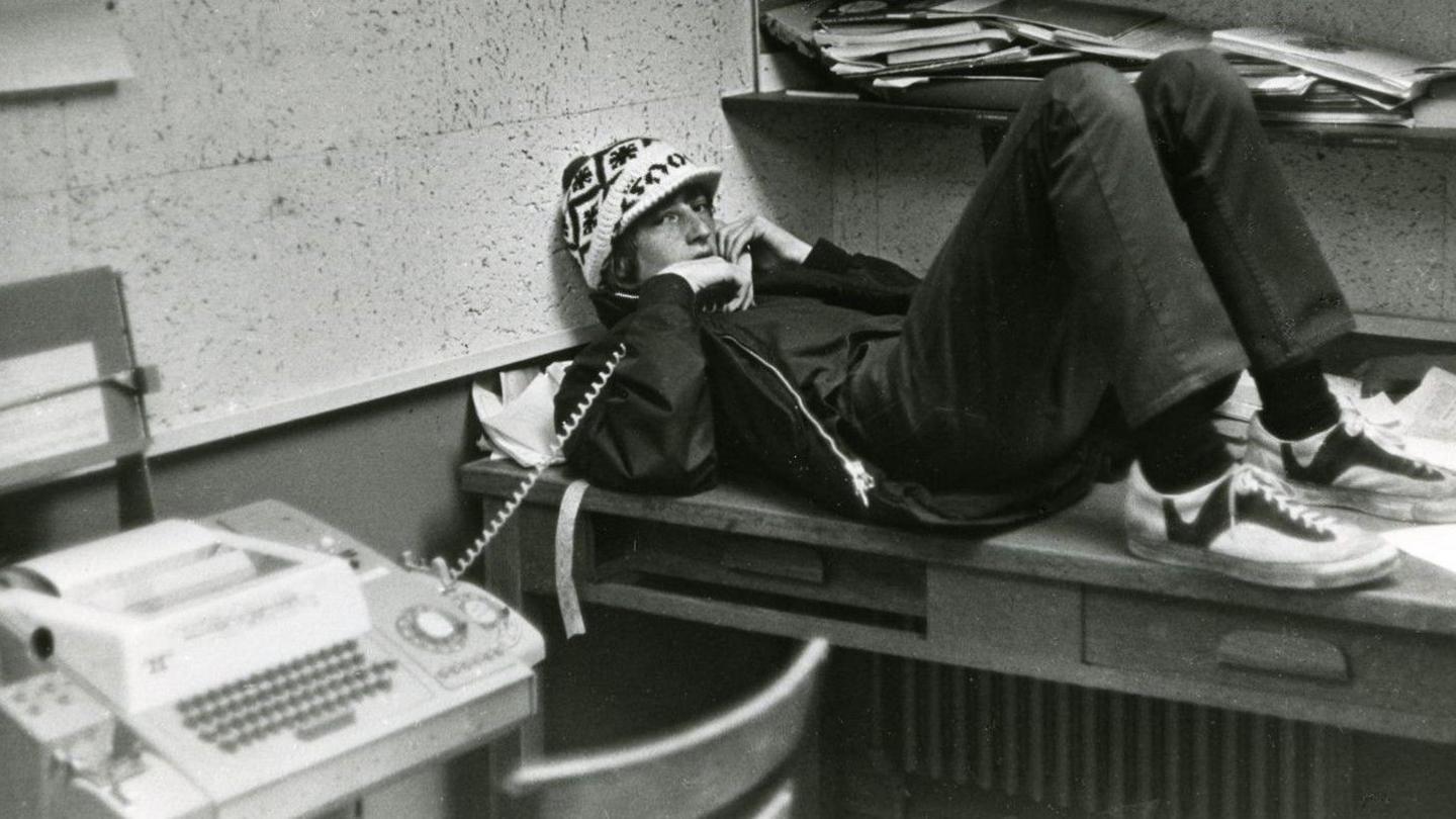 A teenage Bill Gates wearing a hat, lying down on desk, leaning against wall as he holds a landline phone, in a black and white photograph