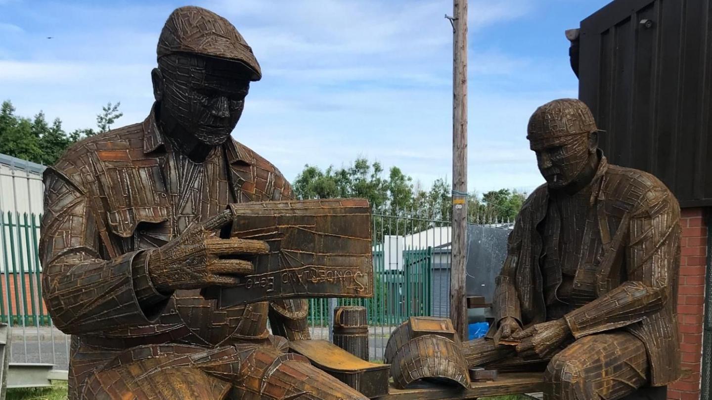 A metal sculpture depicts two men - shipyard workers - sitting on their lunch break while one of them reads the newspaper.