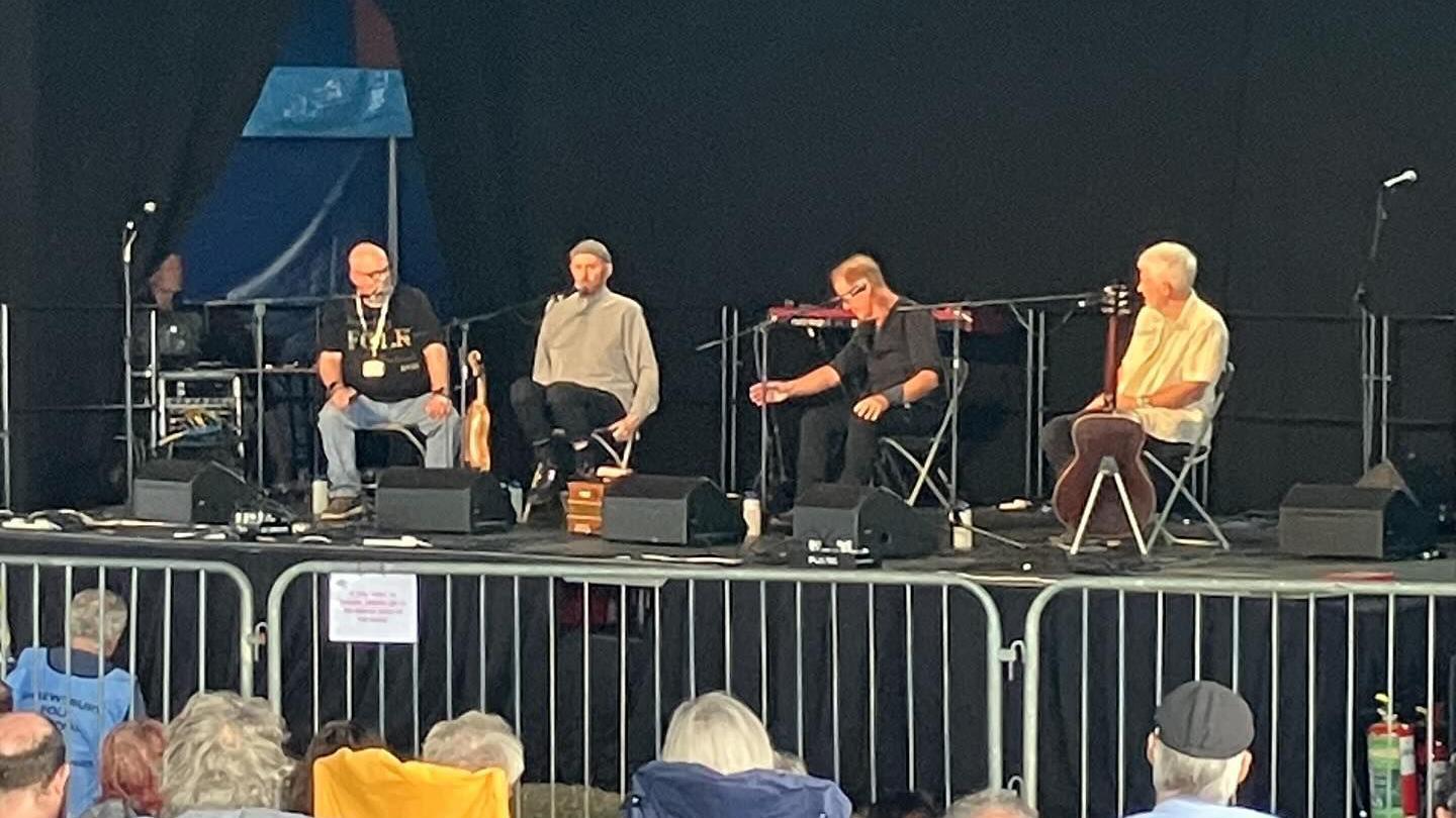 Four men sitting on a stage, delivering a question and answer session to an audience who are sat in front of them on camping chairs at a festival.