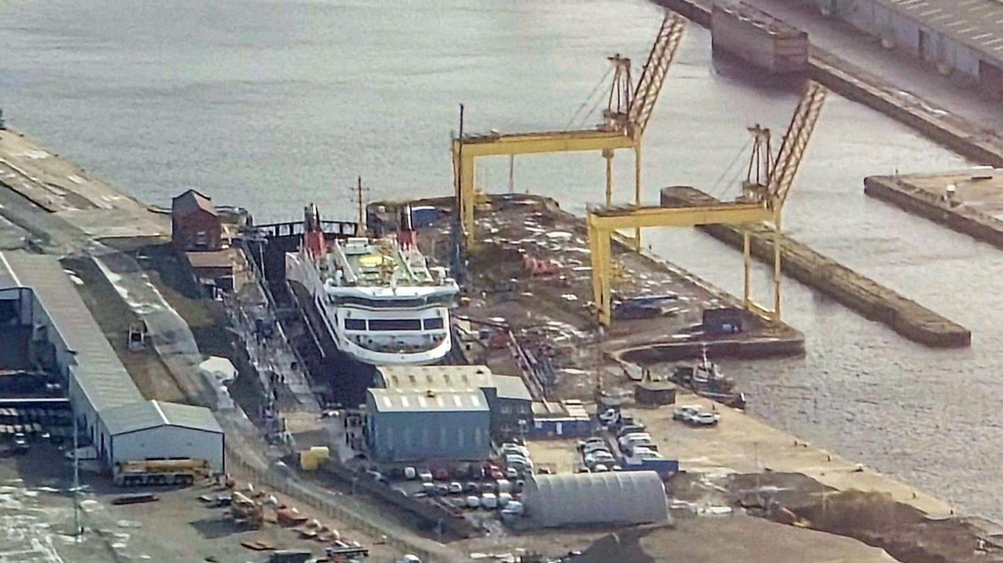 A large ship is seen from an aerial viewpoint in a dry dock with a yellow crane at the side of it