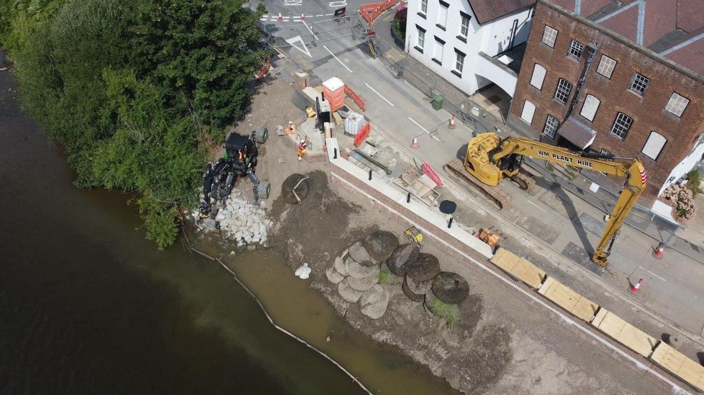 A large yellow machine is in the road near where the wall was being built by trees and the river.