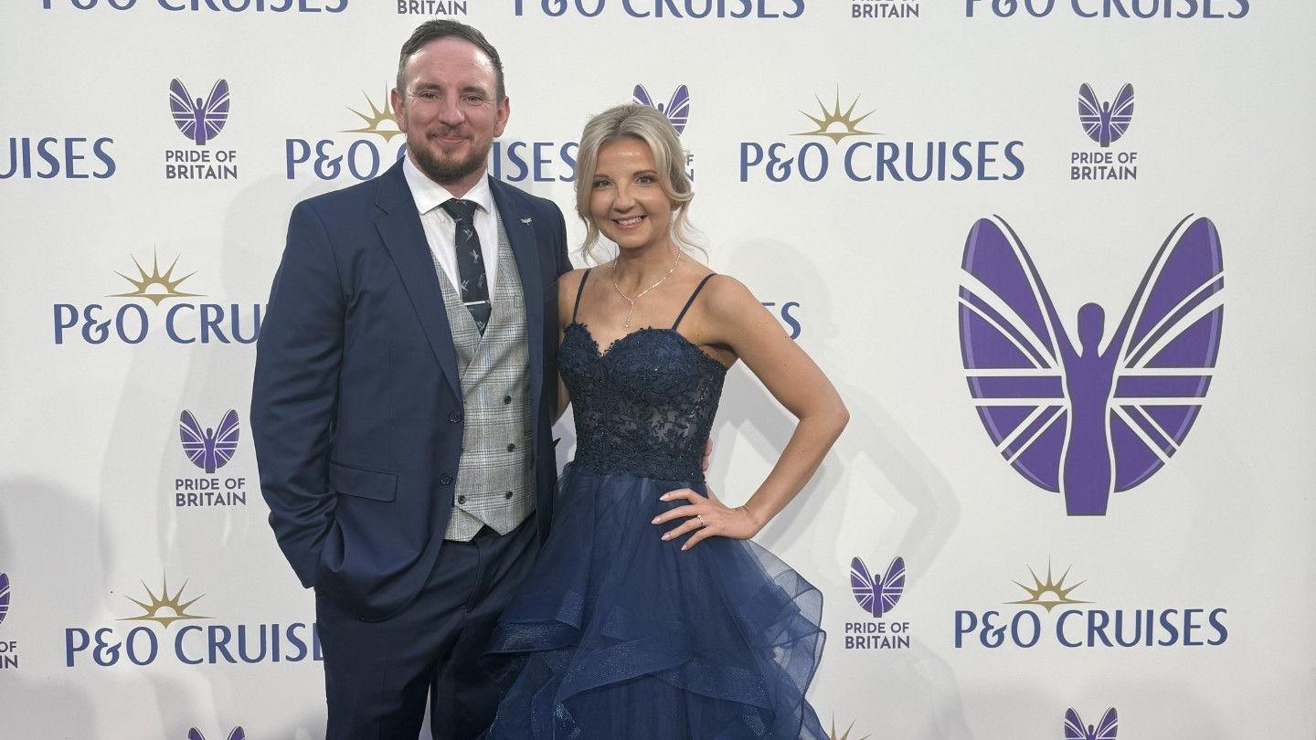 Victoria and Joe Skinner, standing together by a Pride of Britain award sign. He is wearing a blue suit, with blue tie, checked waistcoat, has a beard and short dark hair. Victoria has long blonde hair, tied back and is wearing a long blue ball gown. She has her hand on her hip.