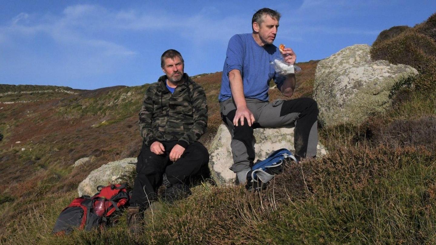 Two men sitting on a hill in Cornwall. One is waring a military-pattern jacket, black trousers and a red backpack. The other is wearing a blue t-shirt and dark grey trousers.