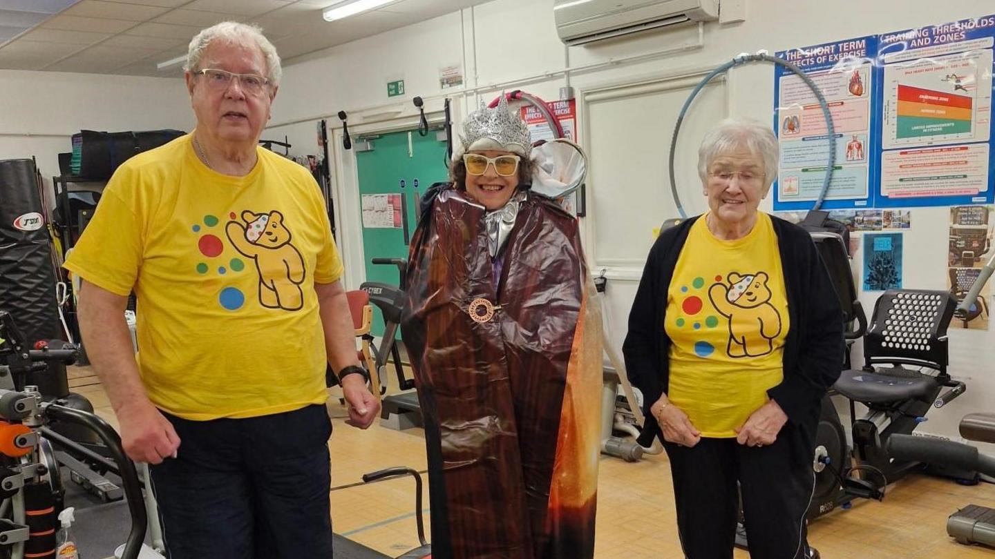 Three people standing in a gym in front of weight lifting machines. The woman in the middle is wearing a fancy dress costume that incorporates The Lion, The Witch and The Wardrobe from the C.S Lewis novel. The two people beside her are wearing yellow Children in Need tops with Pudsey Bear on.
