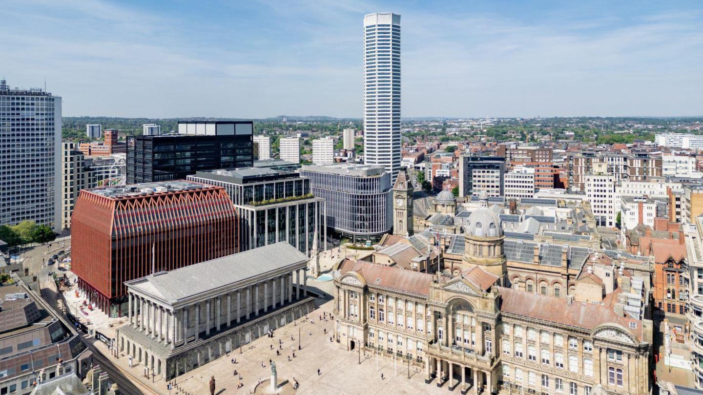 A drone shot of The Octagon towering over Birmingham's Chamberlain Square. It is gleaming white and has eight sides. There is a crown on top. It is a clear day with few clouds in the sky. We can see Birmingham's old-fashioned architecture, including its council house, clashing with the modern design of The Octagon. Because we are higher up we can see even further across the city. There are buildings as far as the eye can see. 