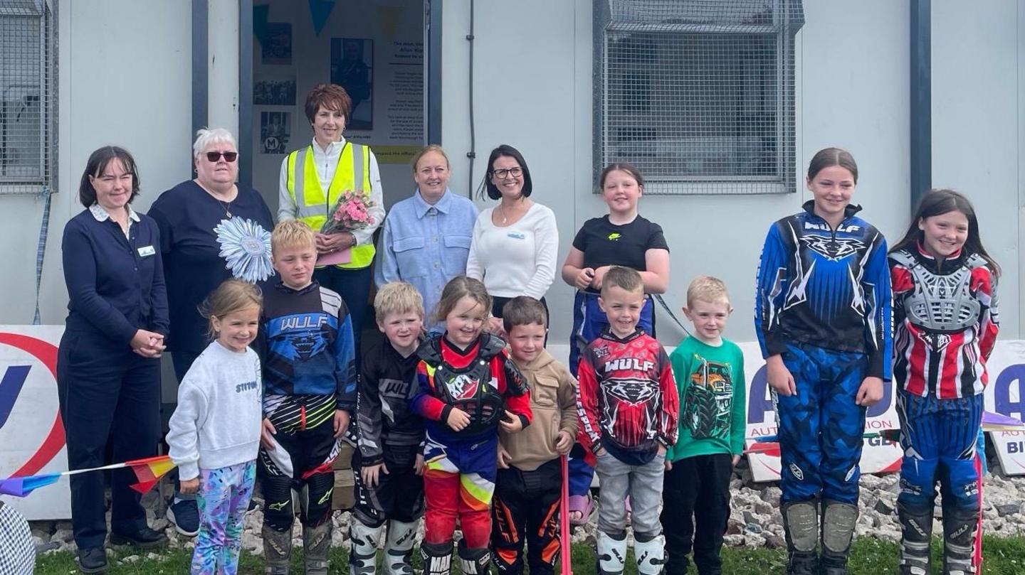 Ten children, some of whom wearing colourful motorbike gear, smile for the cameras. Five adults stand behind them in front of a grey hut.