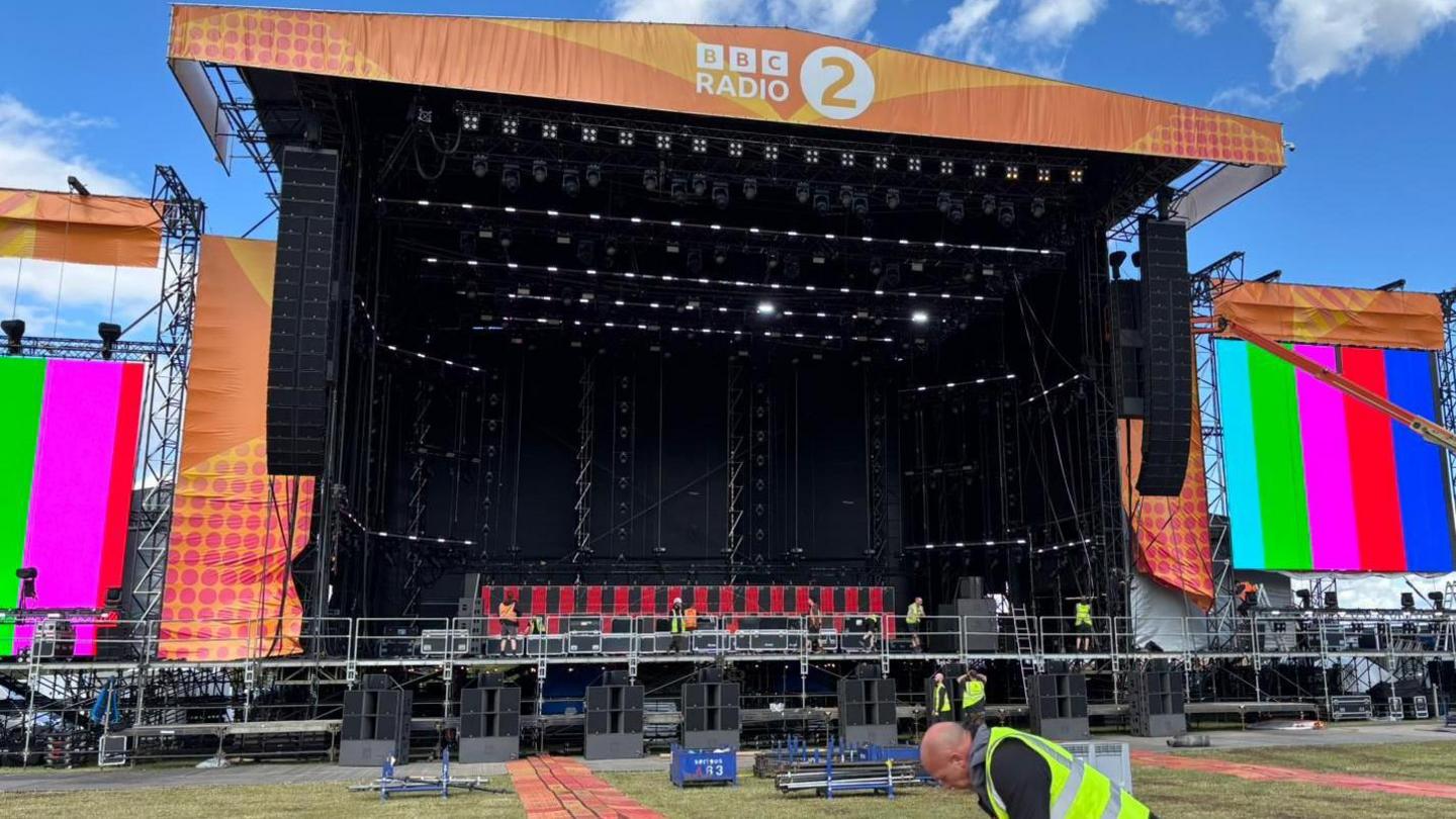 An outdoor music stage with a Radio 2 banner on it and TV screens on the sides. It is being set up, with various workers in hi-vis jackets on stage.