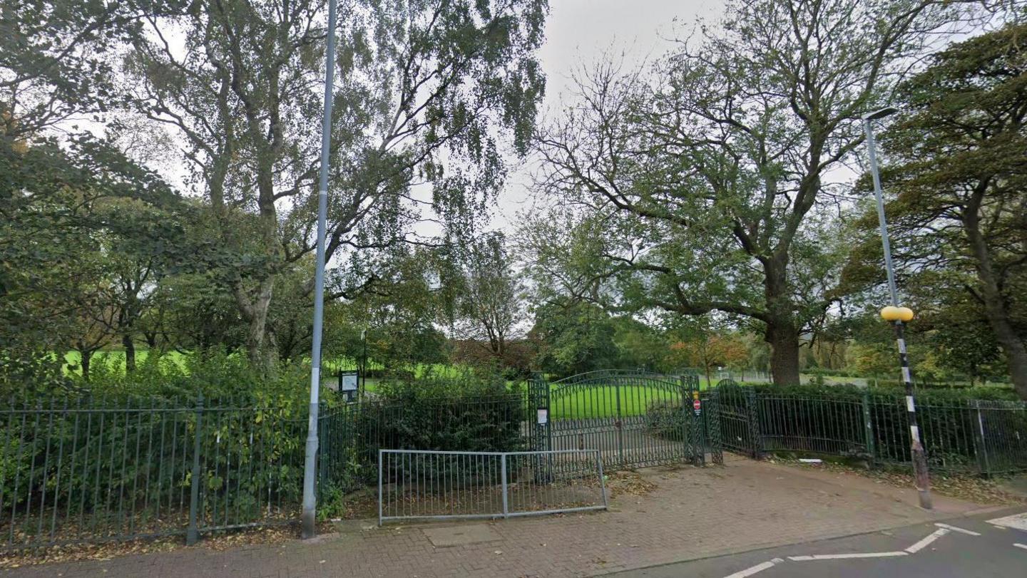One of the entrances to Saltwell Park in Gateshead. A zebra crossing (just in shot on the right hand side) leads to a green pair of gates set into a green metal spiked fence. Behind it is an expanse of green grass and several trees. There is a buckled, grey metal street barrier just outside the gates and a wooden sign to the left. It is an overcast day but the trees are in full leaf.