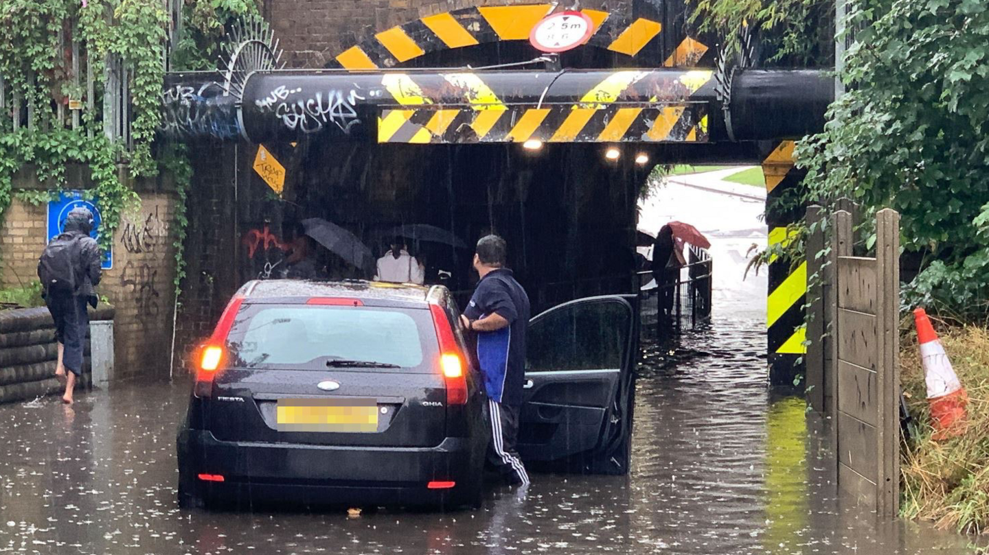 A black Ford Fiesta stuck in floodwater under a low bridge in Slough, with several passers-by in rain coats and holding umbrellas 