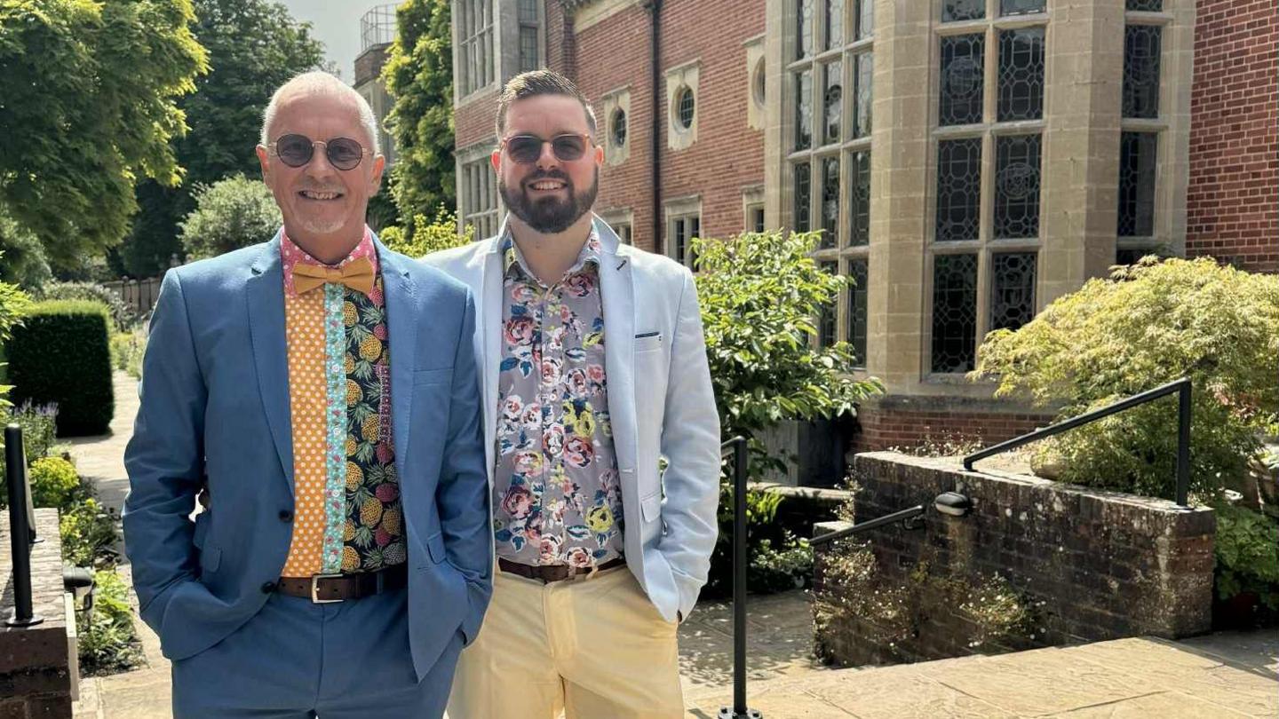 Two men in colourful suits stand smiling in the garden of a stately home. 