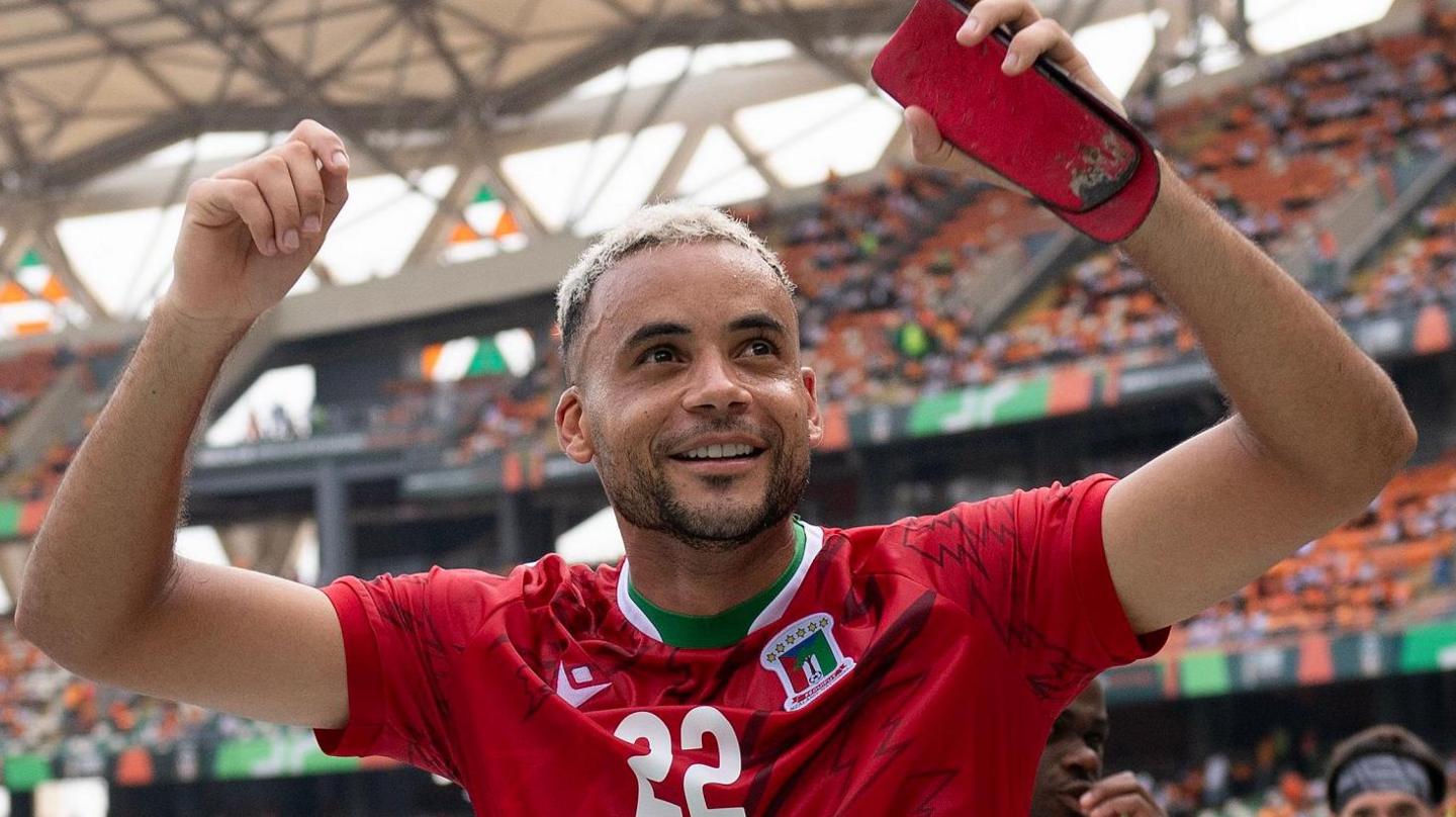 Pablo Ganet, wearing a red Equatorial Guinea shirt with the number 22 on it, pumps both fists towards the crowd while holding a red shin pad in his left hand