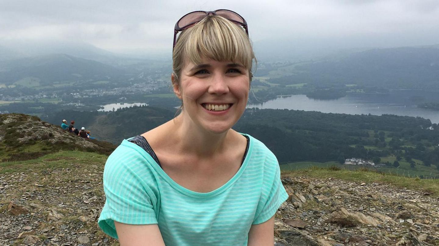 A woman is sitting on top of a mountain in a blue t-shirt. She has blonde hair and is smiling. In the background, there is a landscape of greenery and water. 