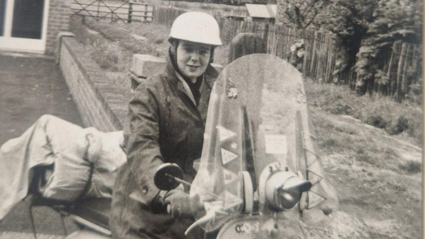 A black and white image of a woman, Margaret Vickers, sitting on a Vespa scooter wearing a crash helmet and gloves.